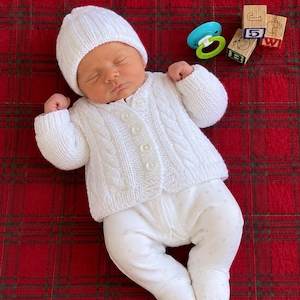 Peut inclure: Un nourrisson endormi portant un bonnet en tricot blanc, un gilet en tricot torsadé blanc avec quatre boutons et un pantalon blanc à pieds. À côté du nourrisson se trouvent une sucette bleue et verte et trois cubes en bois avec J, B, W et 5.
