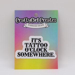 May include: A sticker with the text "IT'S TATTOO O'CLOCK SOMEWHERE" in a white, bold font. The sticker is on a colorful card with the text "CraftyGirl Creates Handmade Sticker". The card has a hole at the top.