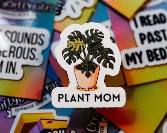 May include: A white sticker with a cartoon illustration of a potted plant with green leaves and a yellow leaf. The text "PLANT MOM" is printed in black below the plant.