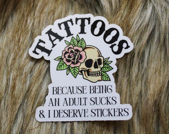 May include: A white sticker with the word "TATTOOS" in black, arched across the top. Below is a skull with a pink rose and green leaves. The text "BECAUSE BEING AN ADULT SUCKS & I DESERVE STICKERS" is printed below the skull.