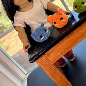 May include: A doll with long black hair sits on a wooden stool with a black top.  The doll is holding three small crocheted stuffed animals: a blue one, an orange one, and a green one.