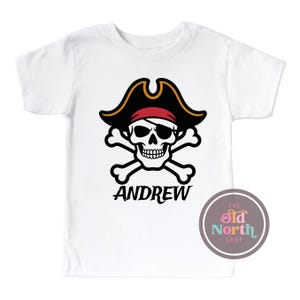 Puede incluir: Camiseta blanca con un dise&ntilde;o de pirata. El dise&ntilde;o presenta una calavera con un sombrero de pirata negro y una bandana roja, con huesos cruzados debajo. El nombre "ANDREW" est&aacute; impreso debajo. El logotipo de The Old North Shop es visible.