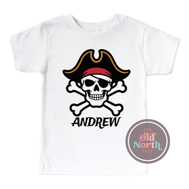 Puede incluir: Camiseta blanca con un dise&ntilde;o de pirata. El dise&ntilde;o presenta una calavera con un sombrero de pirata negro y una bandana roja, con huesos cruzados debajo. El nombre "ANDREW" est&aacute; impreso debajo. El logotipo de The Old North Shop es visible.