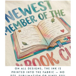 Puede incluir: Primer plano de una tela color crema con una colorida ilustraci&oacute;n de un libro abierto con el texto "NEWEST MEMBER OF THE BOOK CLUB". El libro tiene un dise&ntilde;o de estrella, hoja y coraz&oacute;n. El texto en la parte inferior dice: "ON ALL DESIGNS, THE INK IS PRINTED INTO THE FABRIC - NO DTF, SUBLIMATION OR VINYL FEEL."