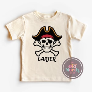 Puede incluir: Camiseta color crema con un dise&ntilde;o de calavera pirata. La calavera lleva un sombrero de pirata negro con ribetes dorados y una bandana roja. Debajo de la calavera est&aacute; el nombre "CARTER" en escritura negra. Un logotipo circular est&aacute; en la esquina inferior derecha.