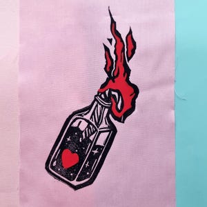 Peut inclure: Impression en linogravure noire et rouge d'une bouteille avec un cœur et des étoiles à l'intérieur, avec des flammes jaillissant du haut. L'impression est sur un tissu rose, sur un fond rose clair et turquoise.