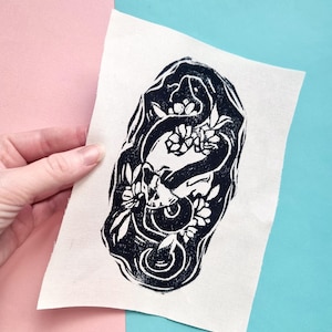 May include: Black and white print of a snake wrapped around a flower on a white fabric patch.