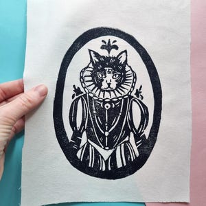 May include: Black and white linocut print of a cat wearing a ruff and a crown, framed in an oval. The cat has three eyes.