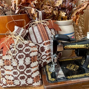 May include: Two decorative fabric pumpkins with brown and cream patterns, tied with raffia and orange ribbon, sit near a vintage black Singer sewing machine. Dried oak leaves and a ceramic jug complete the autumn-themed display.