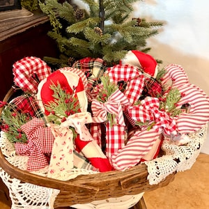 May include: A rustic basket overflowing with festive Christmas decorations. The basket contains red and white candy cane ornaments, adorned with bows and sprigs of greenery. A small evergreen tree sits in the center, adding to the holiday theme.
