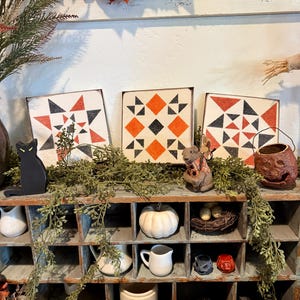 May include: Three square wooden signs with geometric patterns in black, orange, and white. A black cat figurine, a white pumpkin, and a small bird's nest are displayed on a shelf with other decorative items. A carved pumpkin and a fall wreath are also visible.