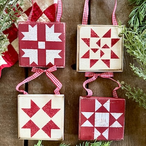 May include: Four square wooden ornaments with red and white quilt block designs. Each ornament has a red and white gingham ribbon for hanging. The ornaments are displayed against a rustic wooden background, with greenery and a quilt in the background.