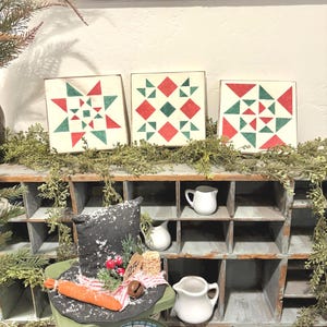 May include: Three square wooden blocks with red and green geometric designs. A black top hat with a carrot, berries, and a bell sits on a green scale. White pitchers are on a wooden shelf.