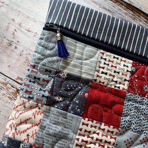 Puede incluir: Un estuche acolchado con cremallera que presenta un dise&ntilde;o de patchwork en telas rojas, blancas y azules. Los patrones incluyen pajaritas peque&ntilde;as, estrellas, motivos florales y texto con capitales de EE. UU. Tiene una cremallera azul con una borla.