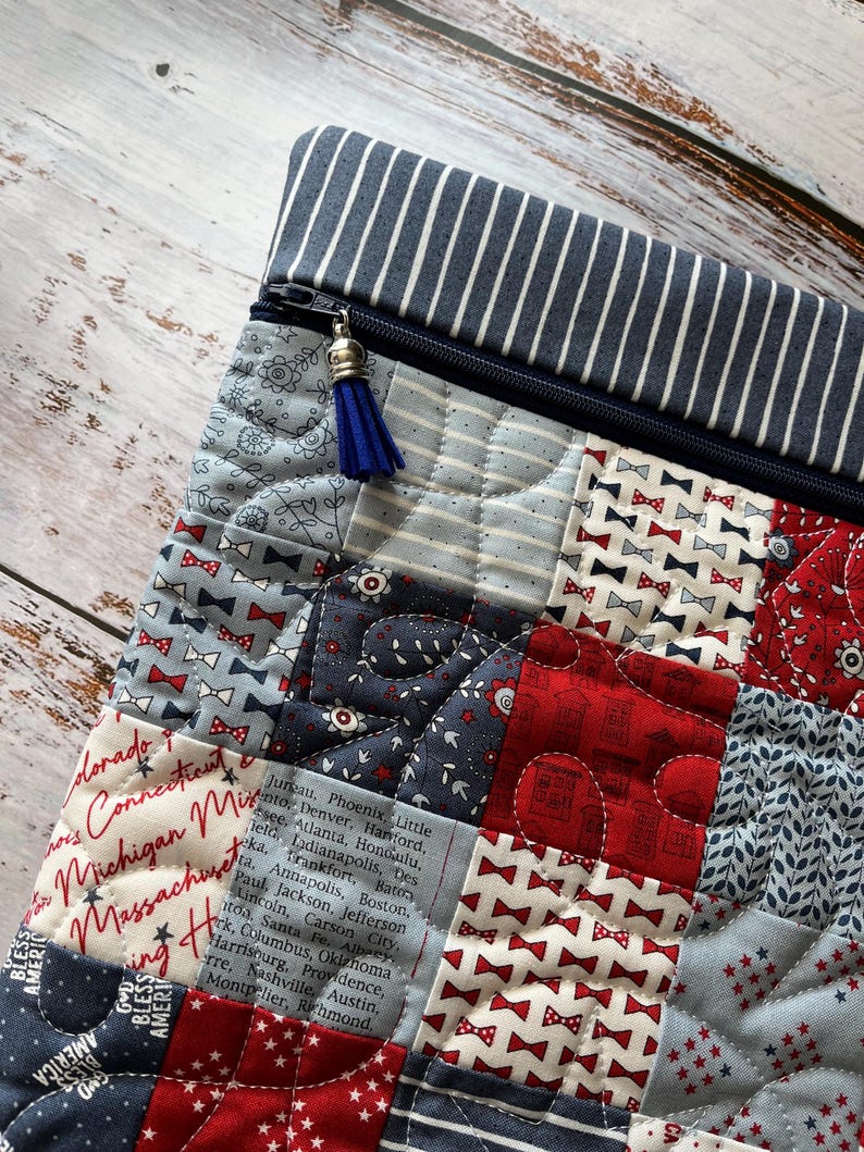 Puede incluir: Un estuche acolchado con cremallera que presenta un dise&ntilde;o de patchwork en telas rojas, blancas y azules. Los patrones incluyen pajaritas peque&ntilde;as, estrellas, motivos florales y texto con capitales de EE. UU. Tiene una cremallera azul con una borla.