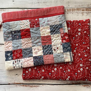 May include: A quilted patchwork zippered pouch with a red and white striped top and a red tassel zipper pull. The patchwork features squares of red, white, and blue patterned fabric. A matching red floral print fabric is next to the pouch.