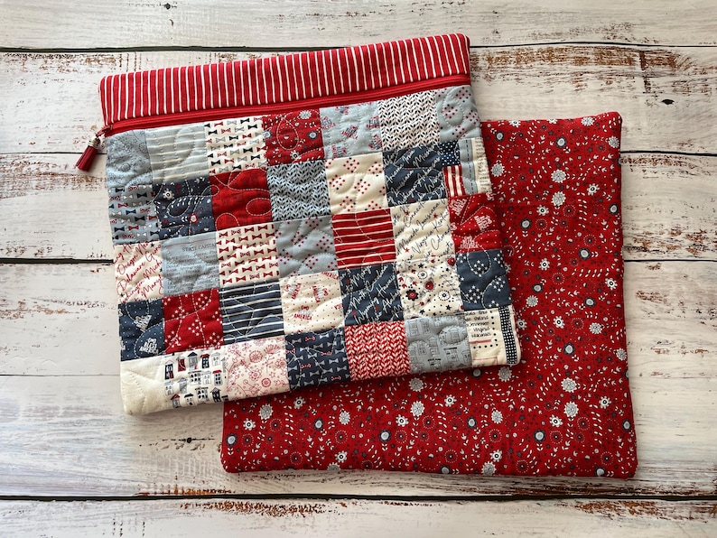 May include: A quilted patchwork zippered pouch with a red and white striped top and a red tassel zipper pull. The patchwork features squares of red, white, and blue patterned fabric. A matching red floral print fabric is next to the pouch.