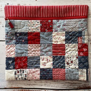 May include: A quilted laptop sleeve with a patchwork design in red, white, and blue. The top features a red and white striped fabric, a red zipper, and a tassel. The patchwork includes various patterns like bows, stripes, and stars.