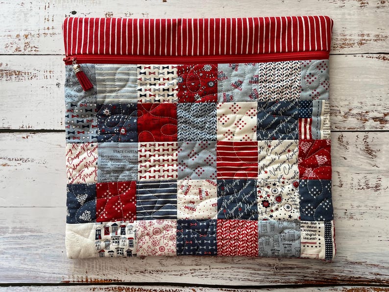 May include: A quilted laptop sleeve with a patchwork design in red, white, and blue. The top features a red and white striped fabric, a red zipper, and a tassel. The patchwork includes various patterns like bows, stripes, and stars.