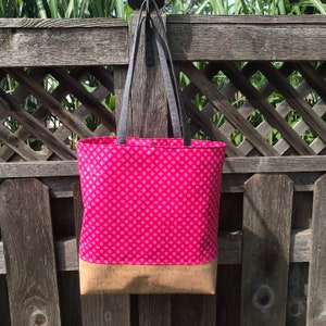 May include: A pink and white patterned tote bag with a cork bottom. The bag has two black straps and is hanging on a wooden fence.