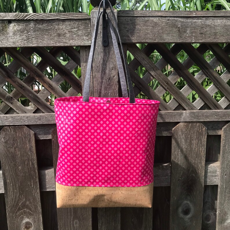 May include: A pink and white patterned tote bag with a cork bottom. The bag has two black straps and is hanging on a wooden fence.