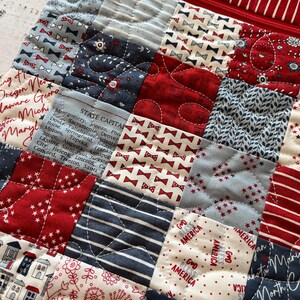 May include: A quilted patchwork bag with a red zipper and a variety of patriotic patterns. The squares feature bow ties, stars, stripes, floral designs, and text including state capitals and "God Bless America."