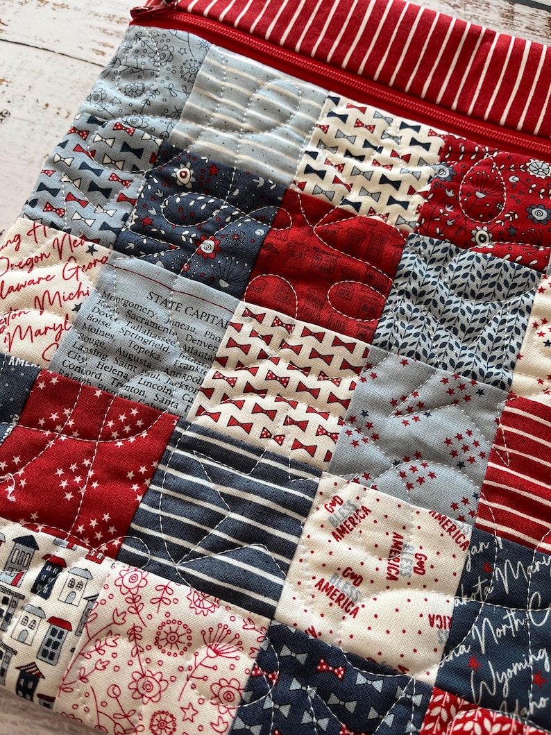 May include: A quilted patchwork bag with a red zipper and a variety of patriotic patterns. The squares feature bow ties, stars, stripes, floral designs, and text including state capitals and "God Bless America."