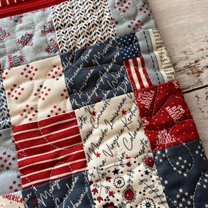 May include: Close-up of a quilted fabric with a patchwork design. The fabric features red, white, and blue squares with star and stripe patterns, state names, and the phrase "God Bless America". A red zipper is visible.