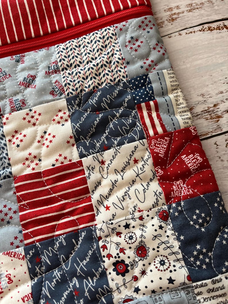 May include: Close-up of a quilted fabric with a patchwork design. The fabric features red, white, and blue squares with star and stripe patterns, state names, and the phrase "God Bless America". A red zipper is visible.