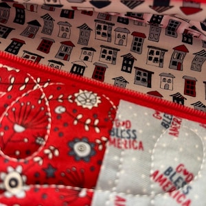 May include: Close-up of a red zippered pouch with a quilted design. The interior lining features a pattern of small houses in various colors. The exterior has a red and white floral pattern, with the text "GOD BLESS AMERICA" visible.