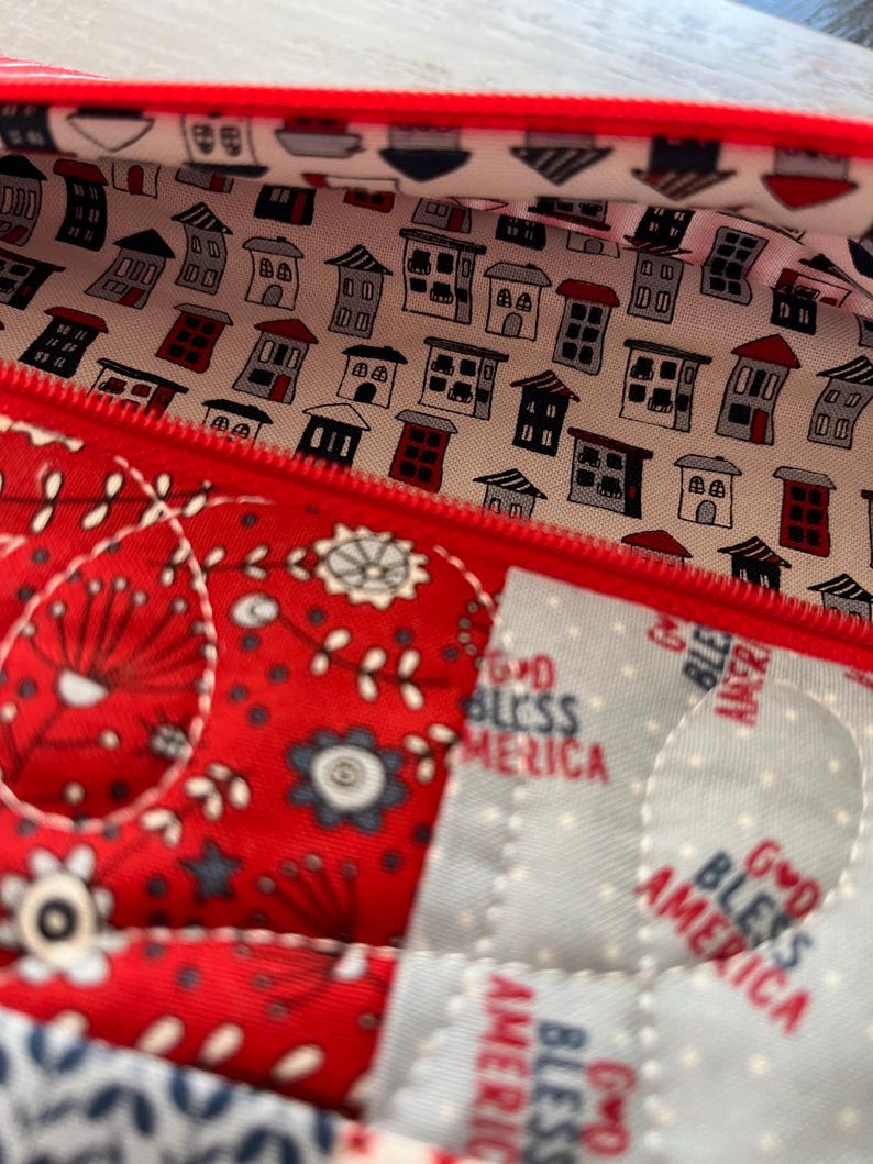 May include: Close-up of a red zippered pouch with a quilted design. The interior lining features a pattern of small houses in various colors. The exterior has a red and white floral pattern, with the text "GOD BLESS AMERICA" visible.