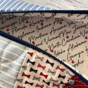 Puede incluir: Un estuche con cremallera que presenta un exterior de patchwork con rayas azules y blancas, un estampado floral rojo y blanco y un estampado de peque&ntilde;os lazos. El forro interior es de tela color crema con nombres de estados de EE. UU. y estrellas rojas.