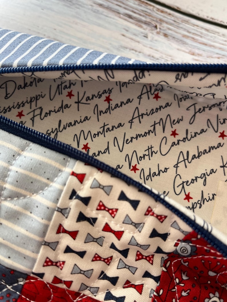 Puede incluir: Un estuche con cremallera que presenta un exterior de patchwork con rayas azules y blancas, un estampado floral rojo y blanco y un estampado de peque&ntilde;os lazos. El forro interior es de tela color crema con nombres de estados de EE. UU. y estrellas rojas.
