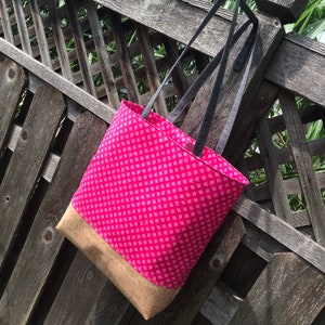May include: A pink tote bag with a cork bottom and black straps. The bag has a pink and white polka dot pattern.