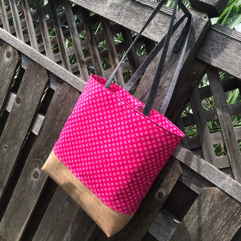 May include: A pink tote bag with a cork bottom and black straps. The bag has a pink and white polka dot pattern.