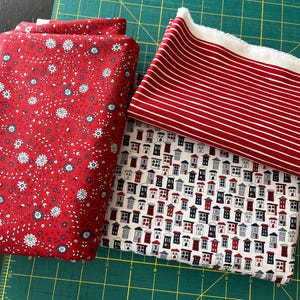 May include: Several fabric pieces in various patterns and colors. One is red with white and blue floral designs, another is red with white stripes, and a third features a repeating pattern of small houses in red, white, and blue. The fabrics are on a green cutting mat.