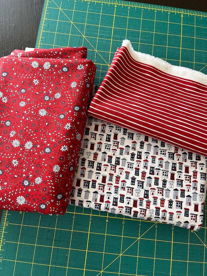 May include: Several fabric pieces in various patterns and colors. One is red with white and blue floral designs, another is red with white stripes, and a third features a repeating pattern of small houses in red, white, and blue. The fabrics are on a green cutting mat.