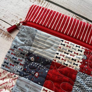 May include: A quilted zippered pouch with a red and white striped top. The pouch features a patchwork design with red, white, and blue fabrics, including bow ties, floral patterns, and text. A red tassel hangs from the zipper.