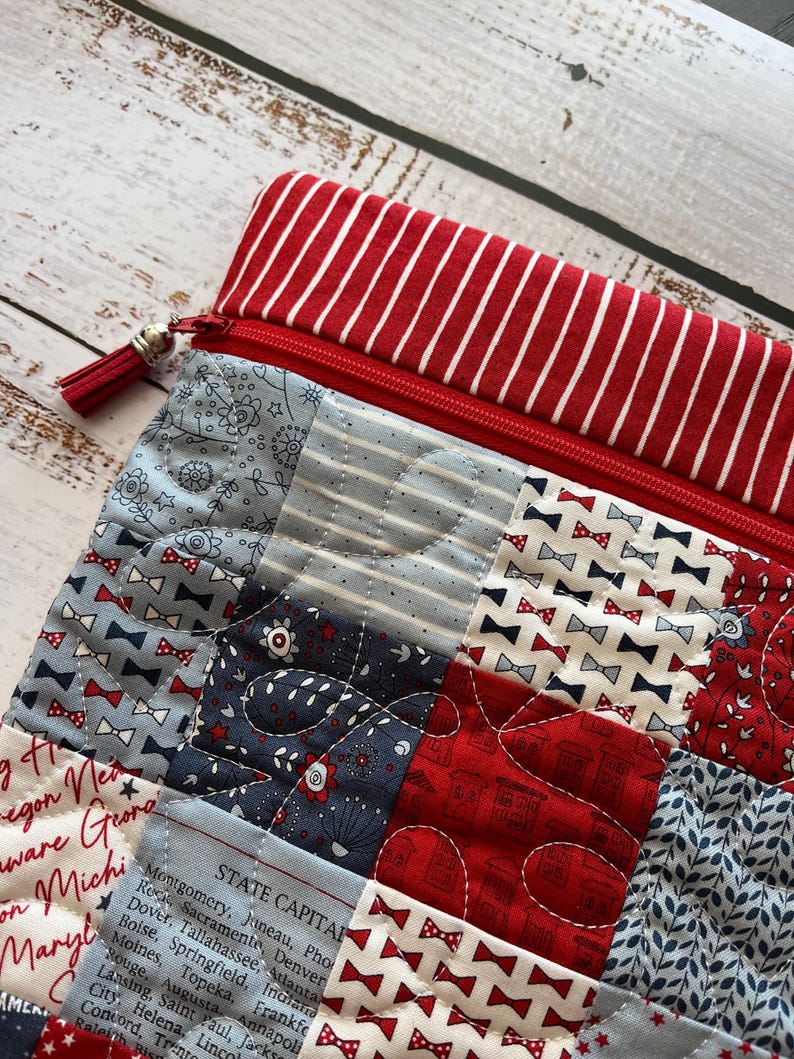 May include: A quilted zippered pouch with a red and white striped top. The pouch features a patchwork design with red, white, and blue fabrics, including bow ties, floral patterns, and text. A red tassel hangs from the zipper.