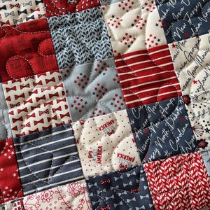 Puede incluir: Una bolsa de tela acolchada con un dise&ntilde;o de patchwork en tonos rojo, blanco y azul. La tela incluye estampados de lazos peque&ntilde;os, rayas, motivos florales y el texto God Bless America junto a nombres de estados de EE. UU. en cursiva.