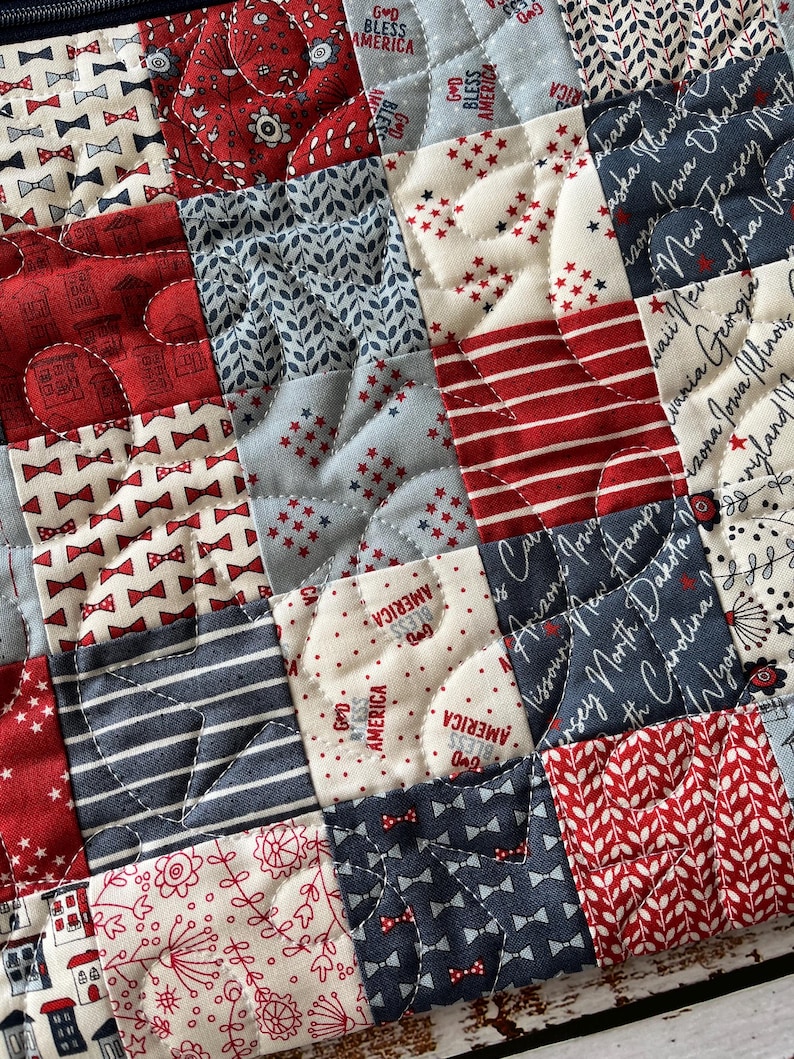Puede incluir: Una bolsa de tela acolchada con un dise&ntilde;o de patchwork en tonos rojo, blanco y azul. La tela incluye estampados de lazos peque&ntilde;os, rayas, motivos florales y el texto God Bless America junto a nombres de estados de EE. UU. en cursiva.