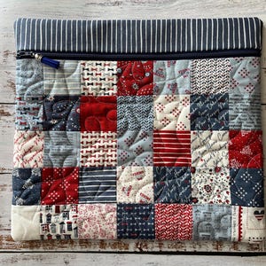 Puede incluir: Un estuche de tela rectangular con un dise&ntilde;o de patchwork de cuadrados rojos, blancos y azules con varios patrones que incluyen estrellas, rayas y texto. La parte superior tiene una cremallera azul oscuro y un borde de tela a rayas.