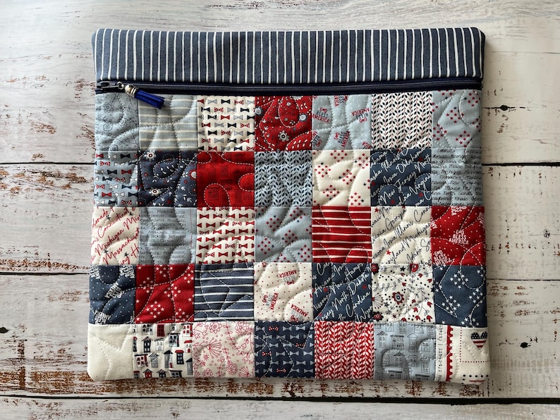 Puede incluir: Un estuche de tela rectangular con un dise&ntilde;o de patchwork de cuadrados rojos, blancos y azules con varios patrones que incluyen estrellas, rayas y texto. La parte superior tiene una cremallera azul oscuro y un borde de tela a rayas.
