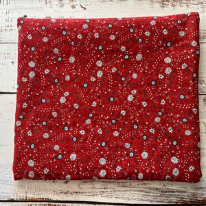 May include: A folded piece of red fabric with a repeating floral pattern. The pattern features small white and blue flowers with dark red and black accents. The fabric is likely cotton or a similar material, suitable for sewing or crafting projects.