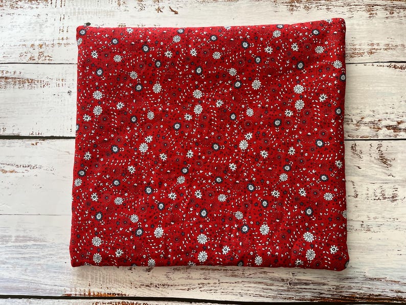 May include: A folded piece of red fabric with a repeating floral pattern. The pattern features small white and blue flowers with dark red and black accents. The fabric is likely cotton or a similar material, suitable for sewing or crafting projects.