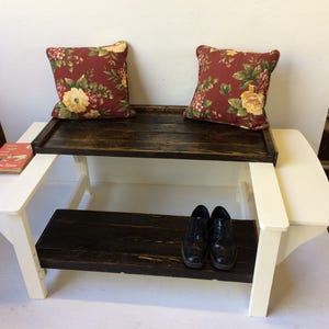 May include: A white wooden bench with a dark brown wooden top and two red floral pillows. The bench has a lower shelf and two pairs of black shoes are on the shelf.