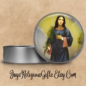 May include: A round metal tin with a lid, featuring a colorful image of a woman holding a branch. The image is surrounded by a clear, glass-like dome. The text "Jays Religious Gifts. Etsy.Com" is printed on the bottom of the image.