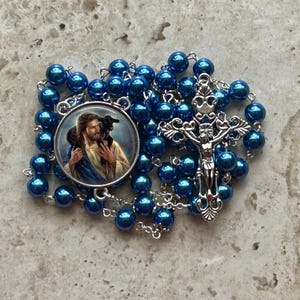 May include: A blue and silver rosary with a round pendant featuring a depiction of Jesus carrying a lamb. The pendant is surrounded by blue beads.