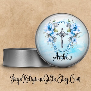 May include: A silver round tin with a blue and white design featuring a cross, a rosary, and blue flowers. The tin has the name "Andrew" printed on it.