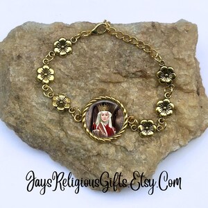 May include: Gold bracelet with a floral design and a round charm featuring a woman in a crown and red robe. The charm is surrounded by a gold frame.